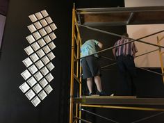 two people standing on scaffolding in front of a black wall with white squares