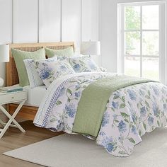 a white bed with blue flowers on the comforter and pillows in front of a window