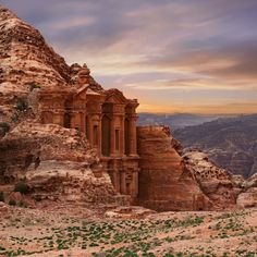 an old building in the middle of some rocks and greenery at sunset with mountains in the background