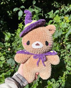 a hand holding a brown teddy bear with a purple hat on it's head