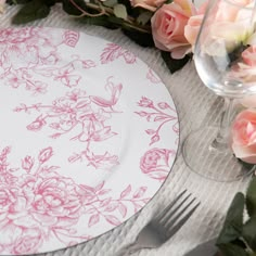 a white plate with pink flowers on it next to a wine glass and some roses