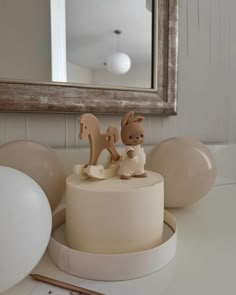 a white cake with an animal figurine on top and balloons in the background