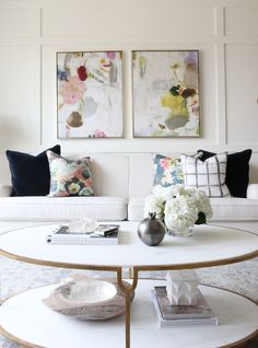 a living room with white couches and two paintings on the wall above it,