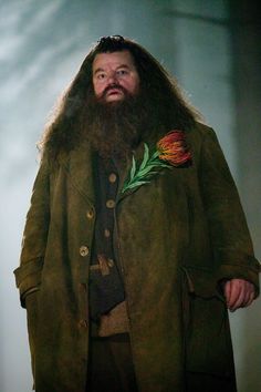 a man with long hair and a beard wearing a green coat is holding a flower in his left hand