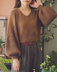 a woman standing in front of a plant wearing a brown sweater and black pants with her hands on her head