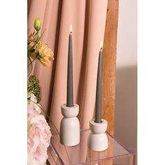 two candles sitting on top of a glass table next to pink flowers and curtained windows