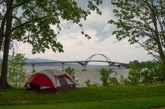 a tent is set up on the grass near trees and water with a bridge in the background