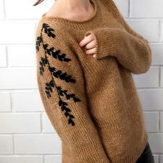 a woman wearing a brown sweater with black leaves on the sleeves and shoulder, standing in front of a brick wall