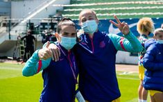 two women in blue jackets and face masks standing on a soccer field with their arms around each other