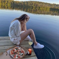 a woman sitting on a dock next to a pizza and drink in front of the water