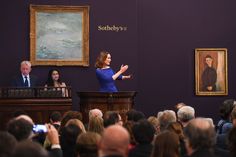 a woman standing at a podium in front of a group of people with paintings on the wall behind her