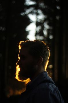 a man standing in the woods at night with his head turned to the side and sunlight shining through trees behind him