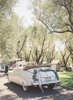 an old white car parked in front of a tree filled driveway with a bow tied to it's roof