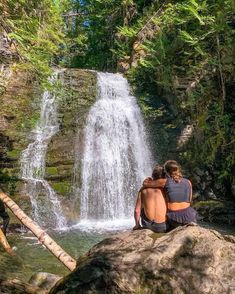 two people sitting on a rock in front of a waterfall