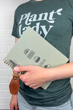 A photo of a woman wearing a dark green shirt that says Plant Lady holding a light green spiral bound journal with plant things on the cover and a brown leather keychain that reads plant lady. The keychain and journal are letter-pressed so they have a slightly indented feel on the design. Plant Lover Shirt Ideas, Plant Lover Tshirt, Plant Lady, House Plants, Green, Plants