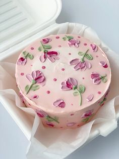 a pink cake with flowers on it sitting in a white box next to a plastic container