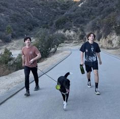 two young men walking their dog down the road in front of some hills and trees