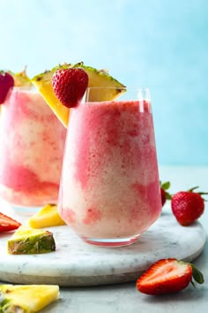 two glasses filled with fruit smoothie next to sliced strawberries and pineapples