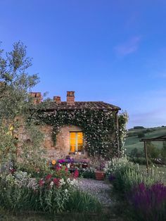 an old stone house surrounded by flowers and greenery in the evening sun, with blue skies above