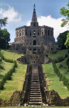 an old building with stairs leading up to it on top of a grassy hill next to trees