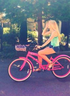 a woman riding a pink bike down a street next to a tree and shrubbery