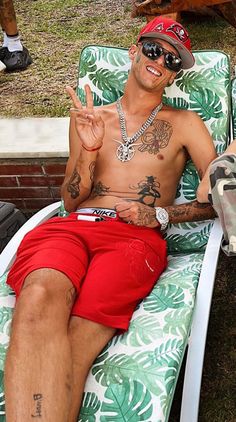 a man sitting in a lawn chair with tattoos on his chest and arms, making the peace sign