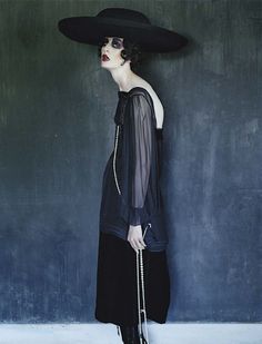 a woman wearing a black hat and dress standing in front of a wall with chains on it