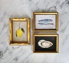 three framed paintings on a marble countertop with fish and lemons in gold frames