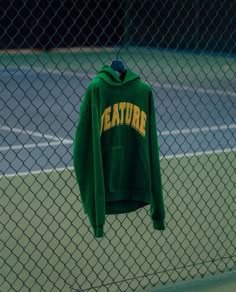 a green hoodie hangs on a chain link fence near a tennis court with the word creature printed on it