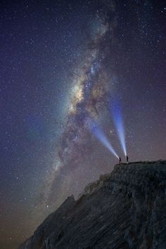 two people standing on top of a mountain under a night sky filled with stars and light beams