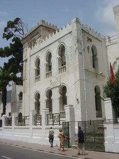 two people are walking down the sidewalk in front of an old white building with arched windows