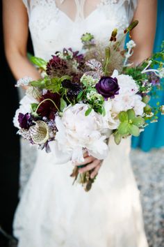 a bride holding a bouquet of white and purple flowers with her groom in the background