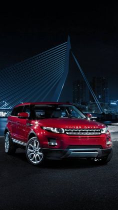 a red range rover parked in front of a bridge at night with city lights behind it