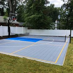 an outdoor basketball court in the middle of a yard with a white fence and trees behind it