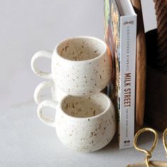 two coffee mugs sitting next to a book and pair of gold - plated scissors