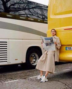 a woman standing in front of a yellow bus reading the paper while looking at her cell phone