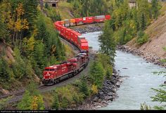 a red train traveling down tracks next to a river and forest covered hillside with trees