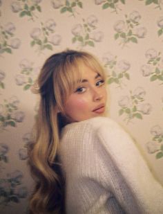 a woman with long blonde hair wearing a white sweater and flowered wallpaper behind her