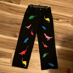 a pair of black pants with colorful dinosaurs painted on the front and back legs, sitting on a wooden floor