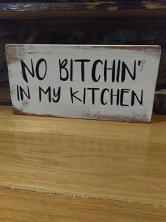 a sign that says no bitchin in my kitchen on a wooden shelf next to a brick wall