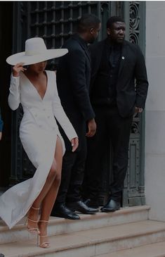 a woman in a white dress and hat is walking down the steps with two men behind her