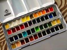 an open box of watercolors on top of a carpeted floor next to a laptop