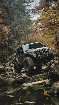 a black jeep driving on rocks in the middle of a stream with fall foliage surrounding it