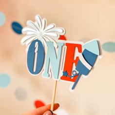 a person holding up a cake topper with the word one in red, white and blue