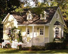 a small yellow house with white picket fence and flowers on the front porch, surrounded by trees