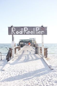 a sign that reads rod reel pier on the beach with people walking along it and bikes parked at the end