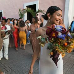 a woman in a white dress holding a bouquet and walking down the street with other people