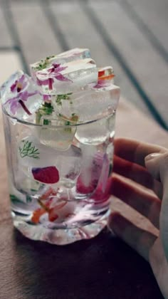 a glass filled with ice and flowers sitting on top of a wooden table next to a person's hand