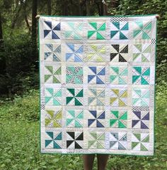 a person holding up a quilt in front of some trees and grass with one block on it's side