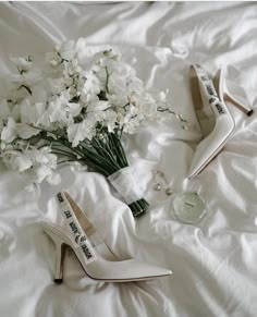 a pair of white high heels sitting on top of a bed next to a bouquet of flowers